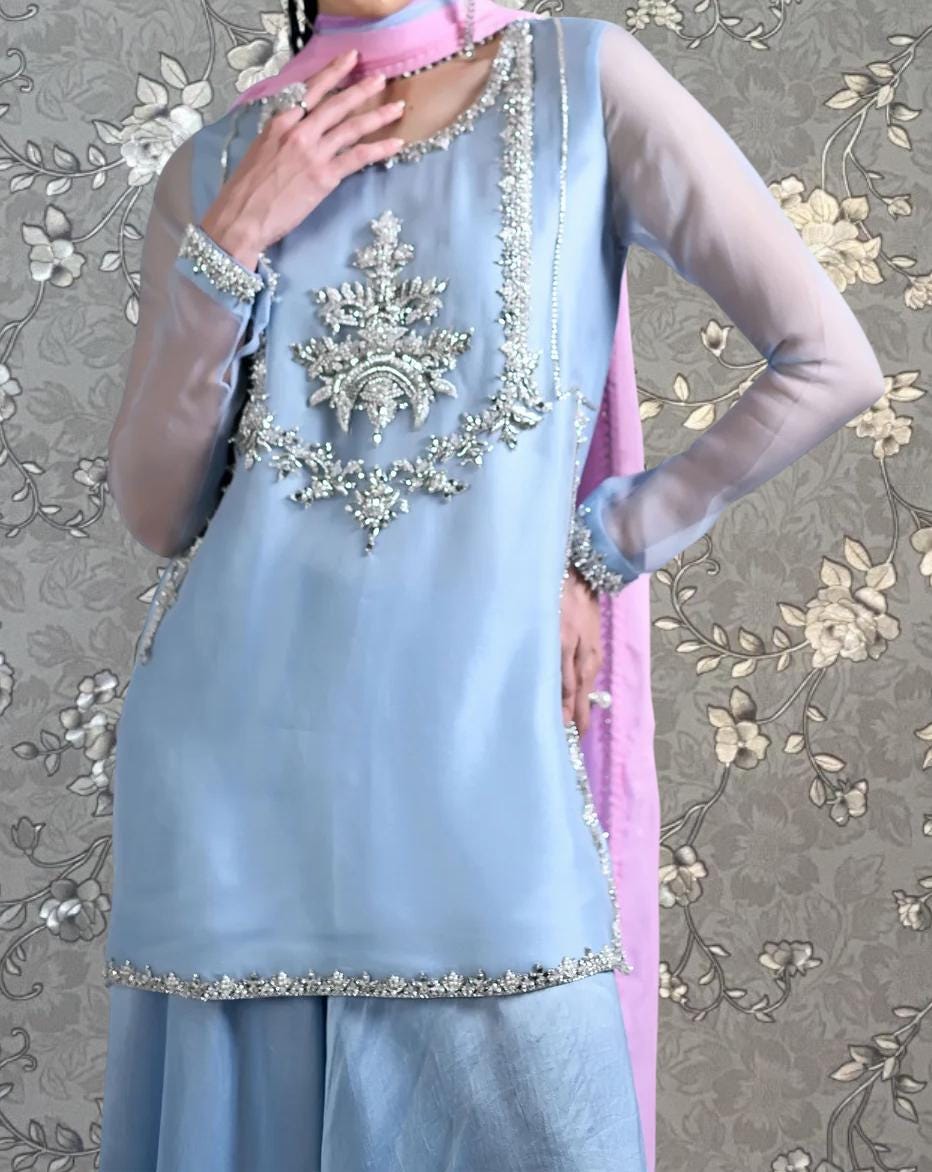 A woman wearing a light blue dress with a pink scarf and a large, intricately designed floral embellishment on the front.