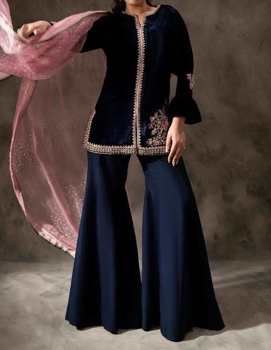 A woman wearing a navy blue dress with gold embroidery and a pink shawl, posing against a gray background.