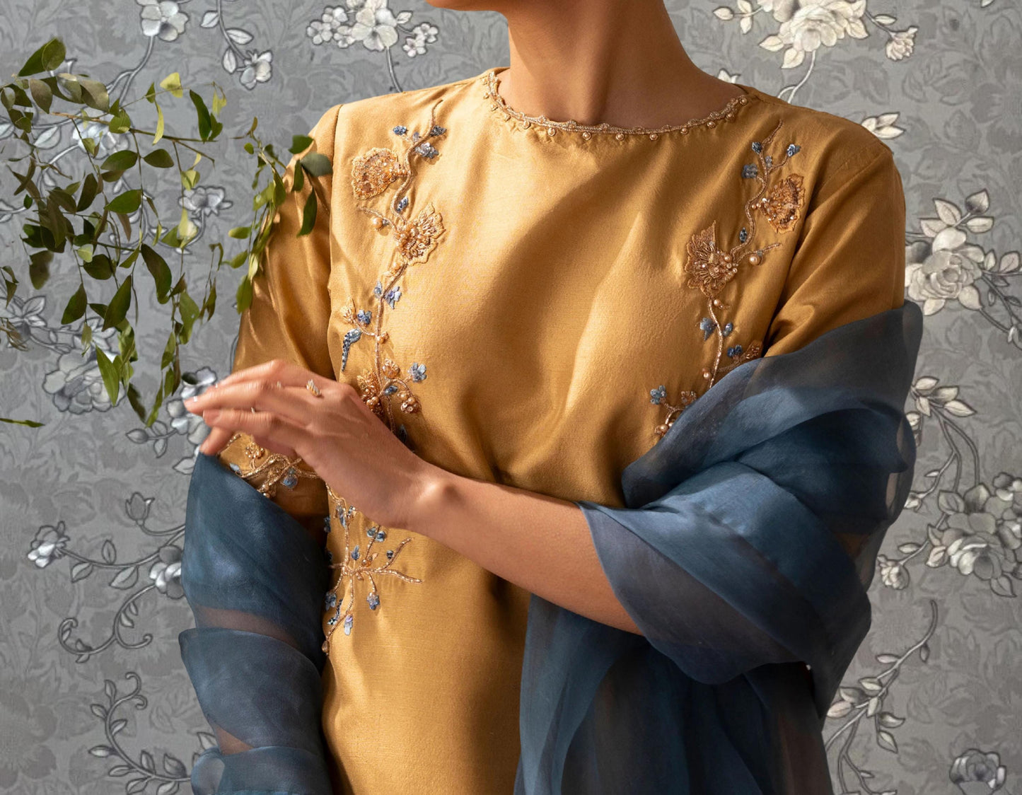 A woman wearing a gold dress with floral embroidery stands in front of a gray floral wallpaper.