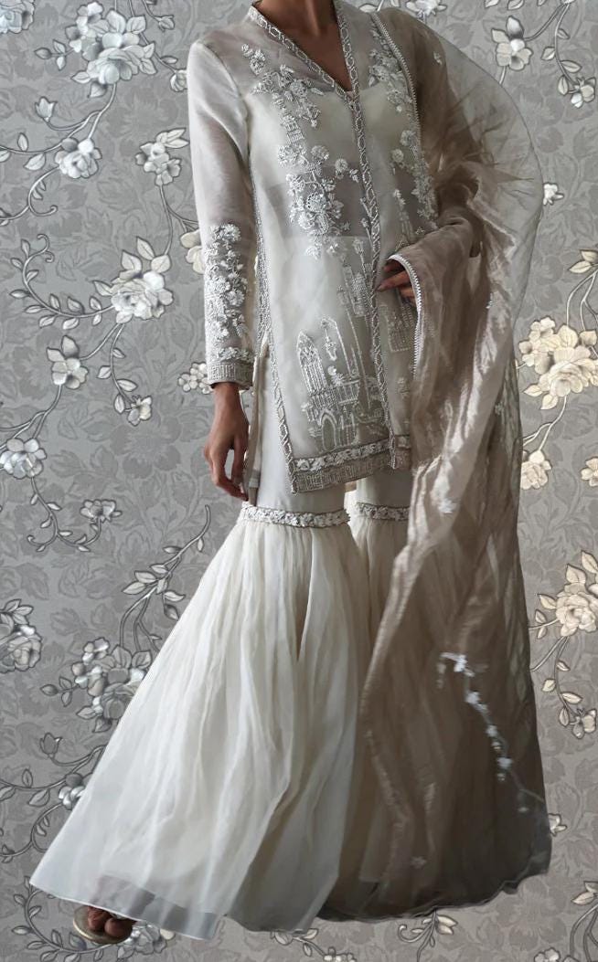 A woman wearing a long, white dress with intricate embroidery stands against a gray floral background.