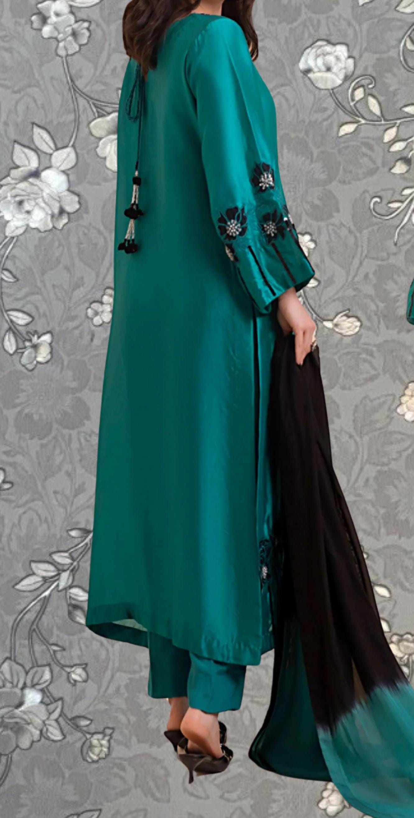 A woman wearing a long, green dress with floral embroidery and holding a black shawl stands against a gray floral background.