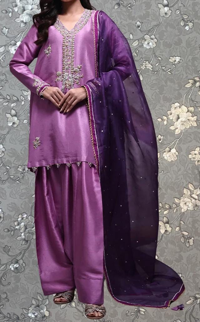 A woman wearing a purple and purple dress with a matching purple shawl, standing against a gray floral background.