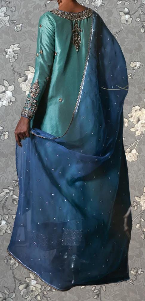 A person wearing a long, green dress with a blue shawl draped over their shoulders, standing against a floral background.