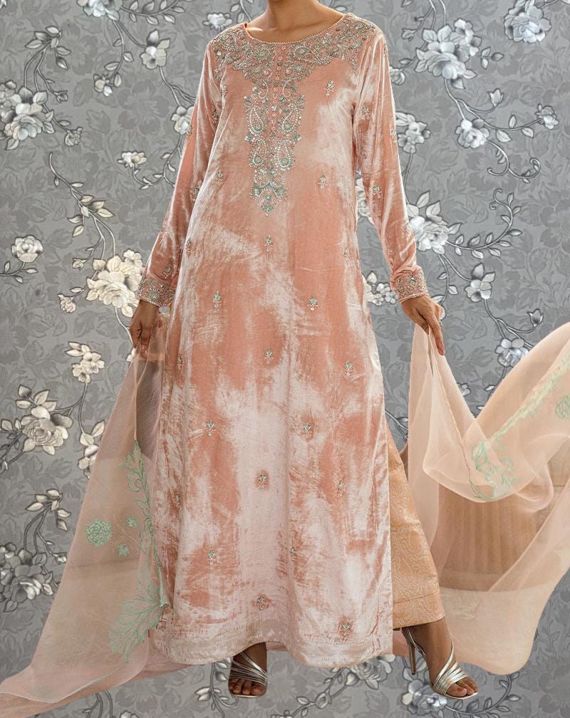 A woman wearing a long, peach-colored dress with intricate embroidery stands against a gray floral background.