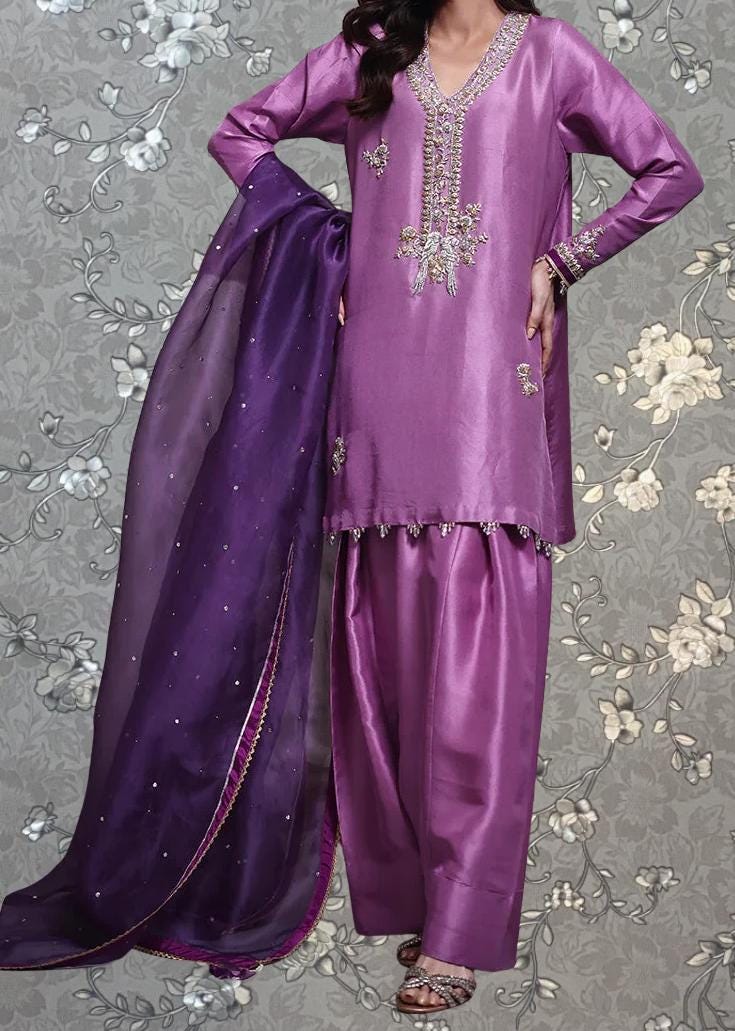 A woman wearing a purple dress with intricate embroidery and a matching purple shawl stands against a gray floral background.