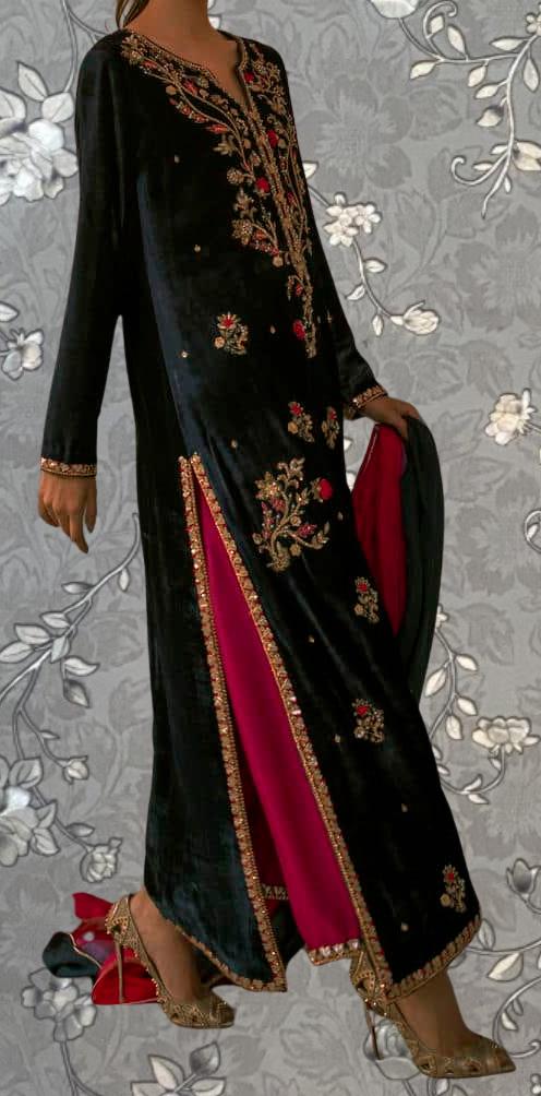 A person wearing a long black dress with gold embroidery and red accents, standing against a gray floral background.