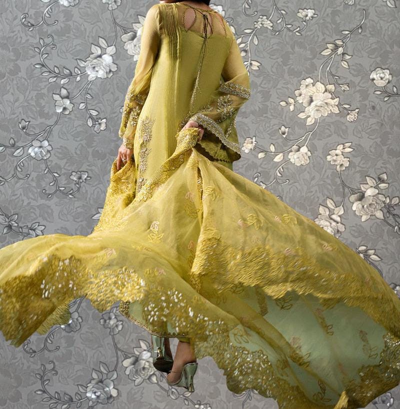 A woman wearing a long, flowing yellow dress with intricate embroidery and floral patterns, standing against a gray floral background.