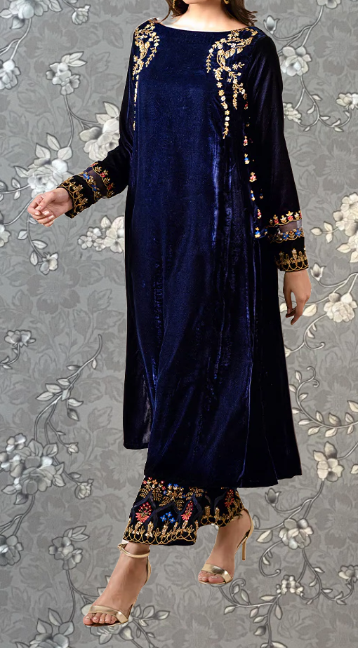 A woman wearing a long navy blue velvet dress with gold embroidery and floral patterns, paired with gold heels, stands against a gray floral background.