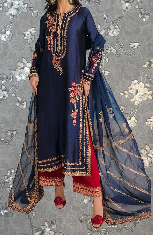 A woman wearing a navy blue dress with gold embroidery and red pants, standing against a gray floral background.