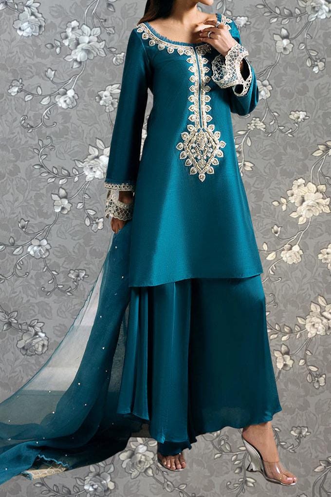 A woman wearing a teal blue dress with intricate white embroidery and a matching skirt, standing against a gray floral background.