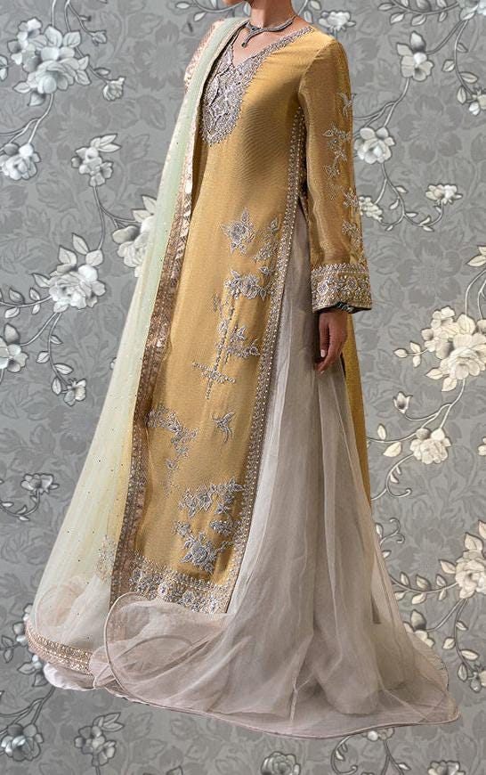 A gold and white dress with intricate embroidery and a long, flowing skirt is displayed against a gray floral background.