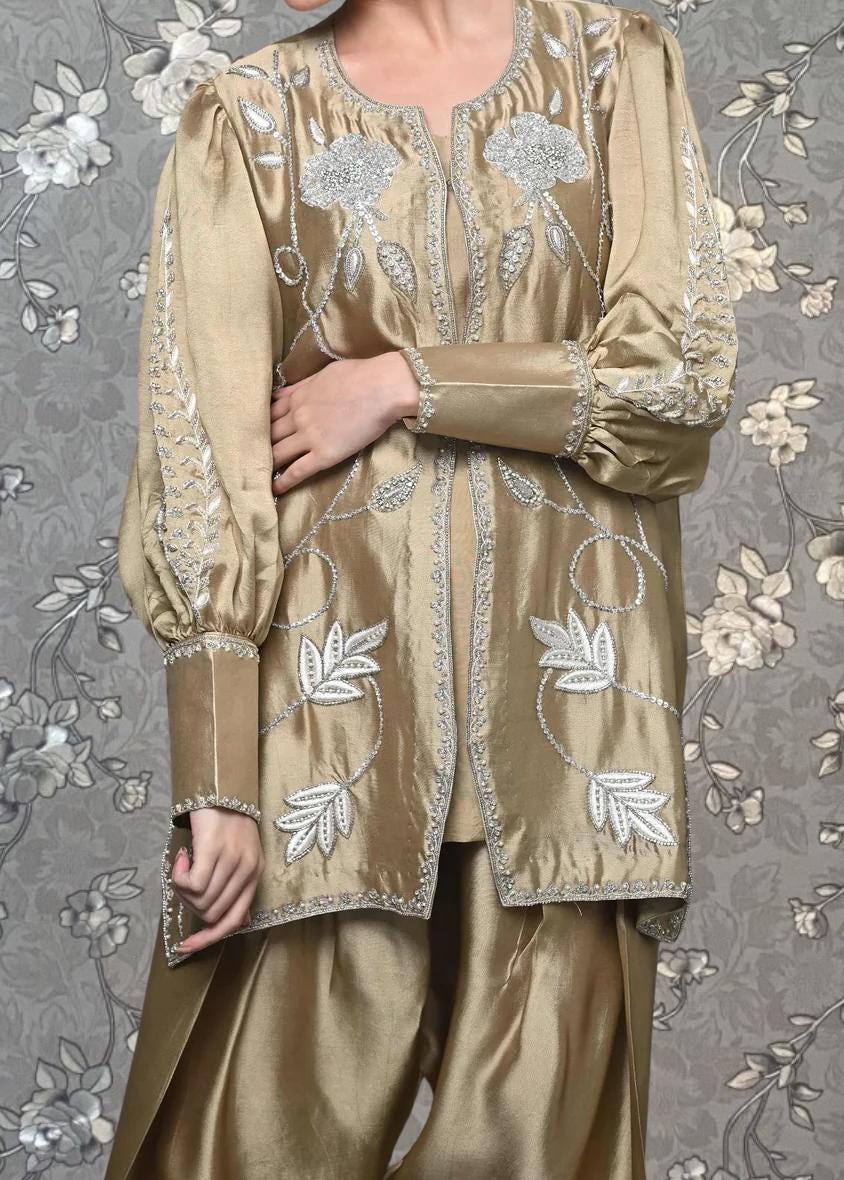 A person wearing a gold-colored dress with intricate embroidered designs, standing against a gray floral background.