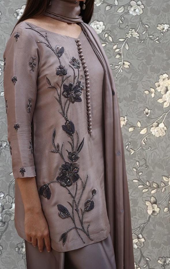 A woman wearing a grey dress with intricate floral embroidery stands against a floral patterned background.