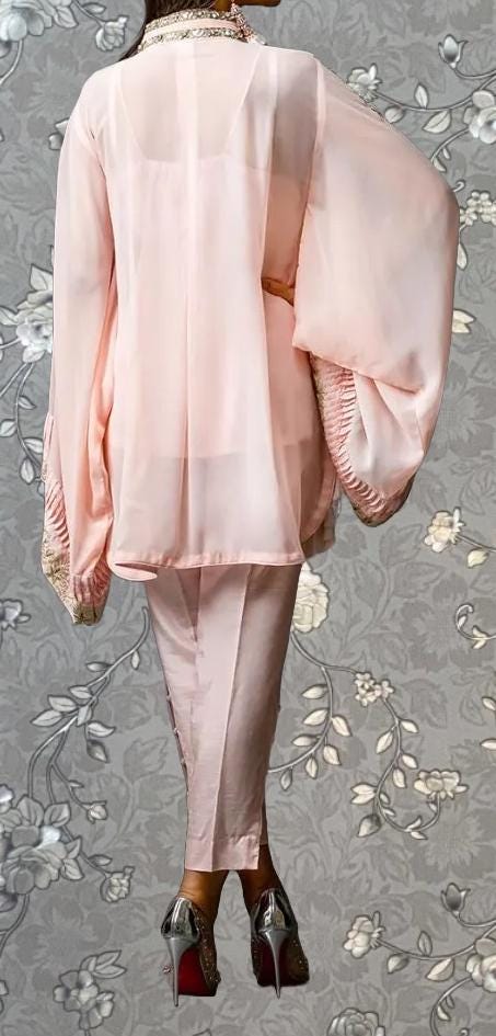 A person wearing a pink outfit with long sleeves and a high collar, standing against a gray floral background.