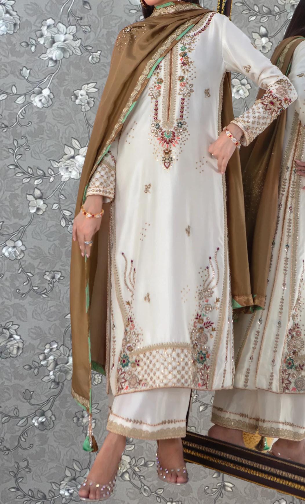 A person wearing a white and gold dress with intricate embroidery and a brown shawl, standing in front of a floral patterned background.