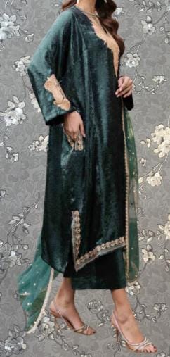 A woman wearing a long, green velvet dress with gold accents, standing against a gray floral background.