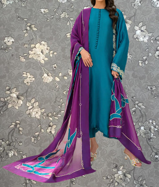 A woman wearing a teal dress with a purple shawl and gold accents, standing against a gray floral background.