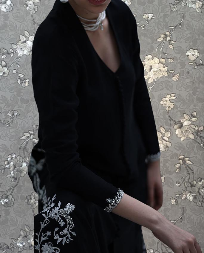 A woman wearing a black dress with a floral pattern and a matching necklace stands against a floral background.