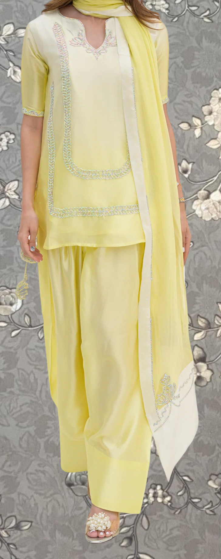 A woman wearing a yellow dress with a matching yellow scarf, standing against a gray floral background.