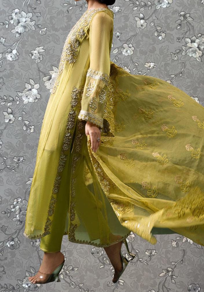 A woman wearing a long, yellow dress with intricate embroidery and a flowing skirt, standing against a gray floral background.