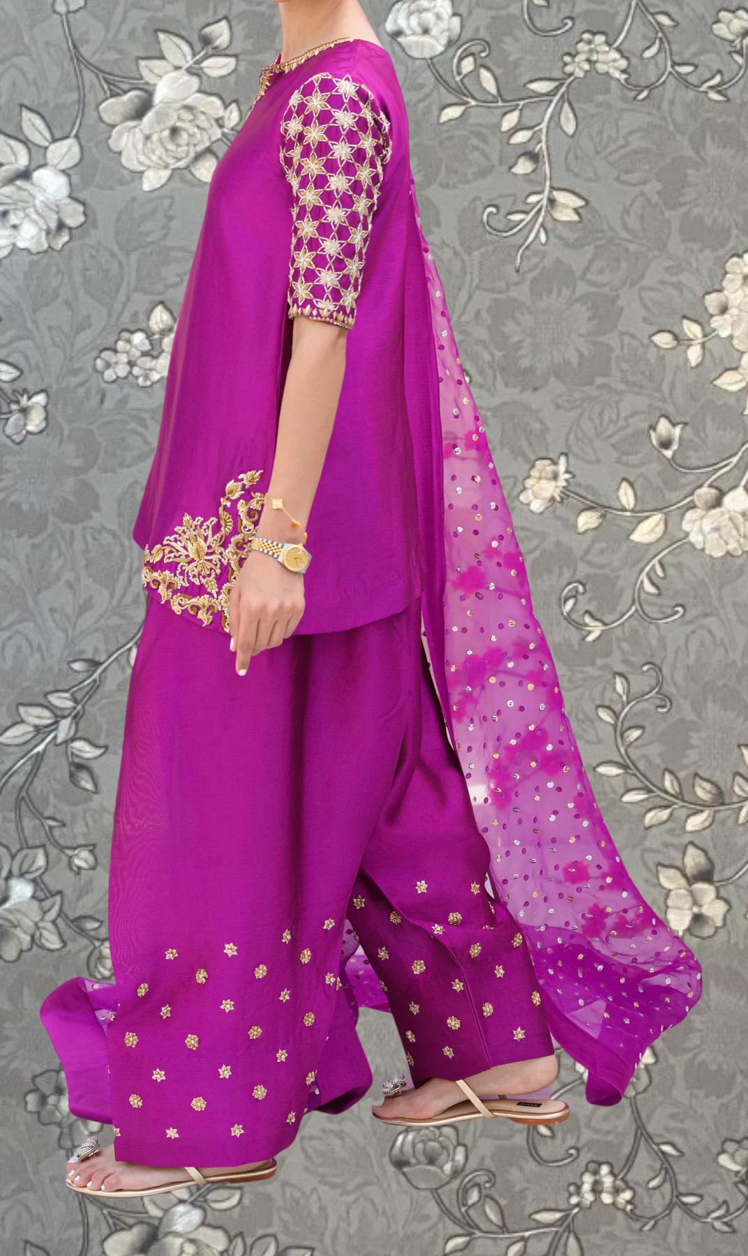 A person wearing a vibrant purple dress with gold embroidery and a matching pink shawl, standing against a gray floral background.