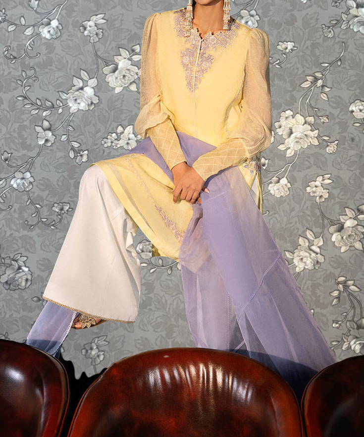 A woman wearing a yellow and purple dress is sitting on a chair in front of a gray floral wallpaper.