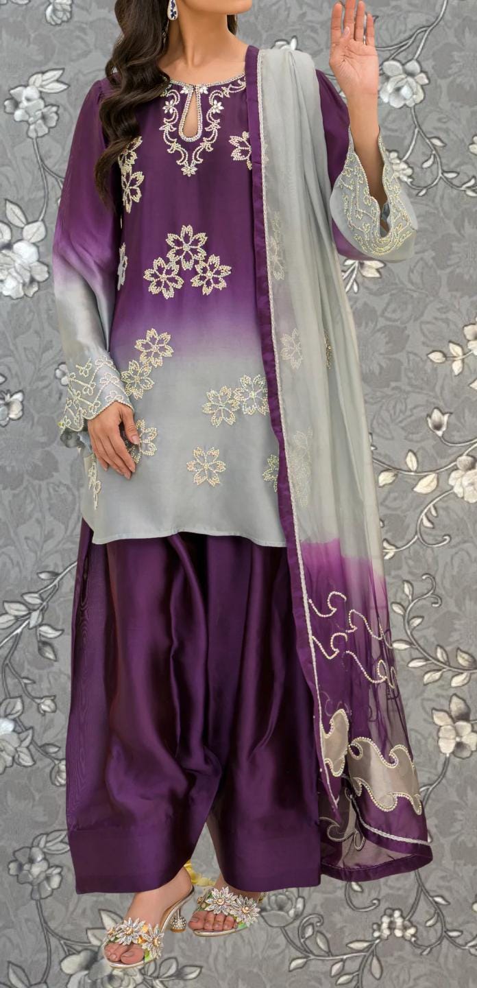 A woman wearing a purple and gray dress with intricate embroidery, standing against a gray floral background.