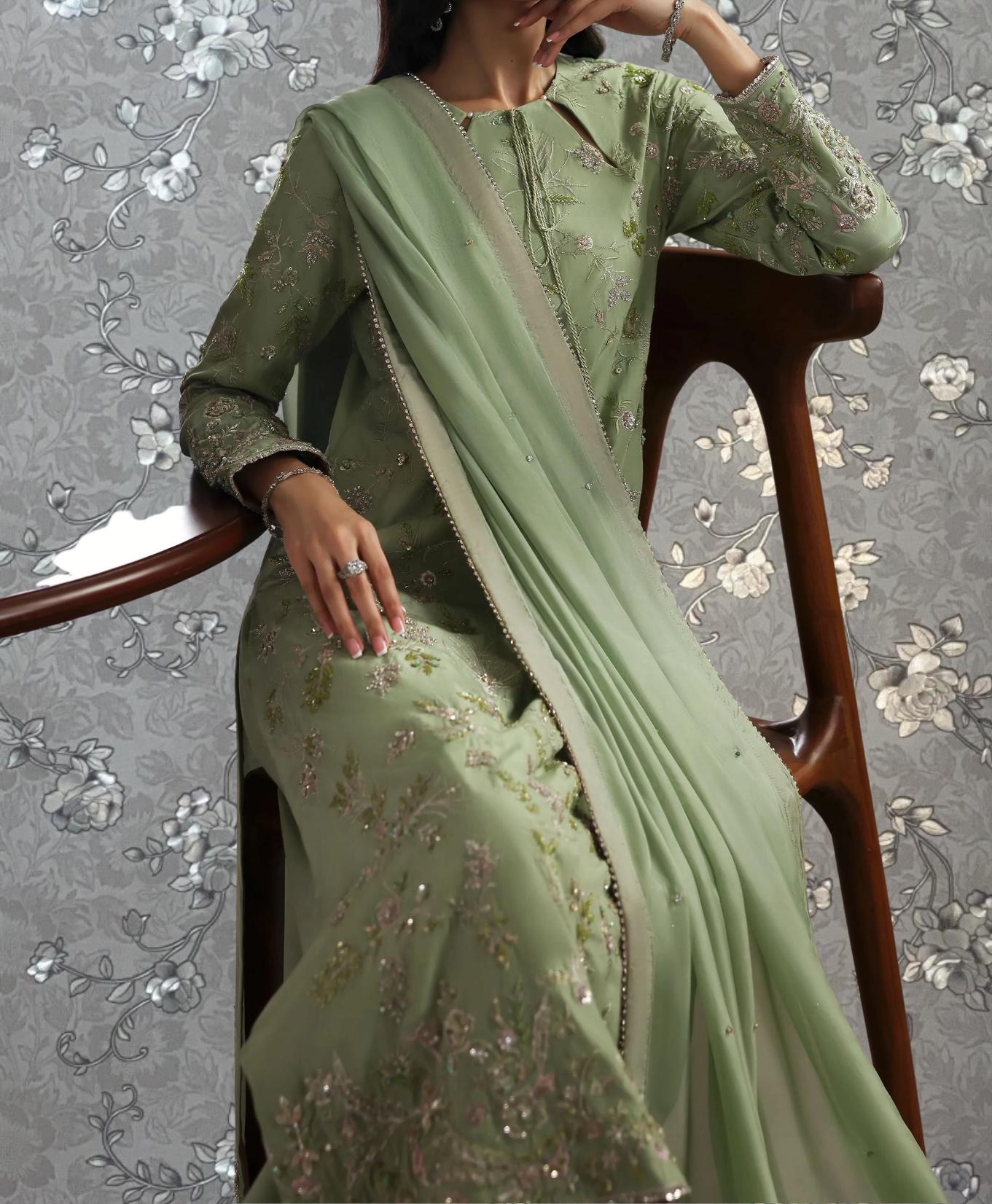 A woman wearing a green dress with intricate embroidery and a matching green shawl is seated on a wooden chair against a floral patterned background.