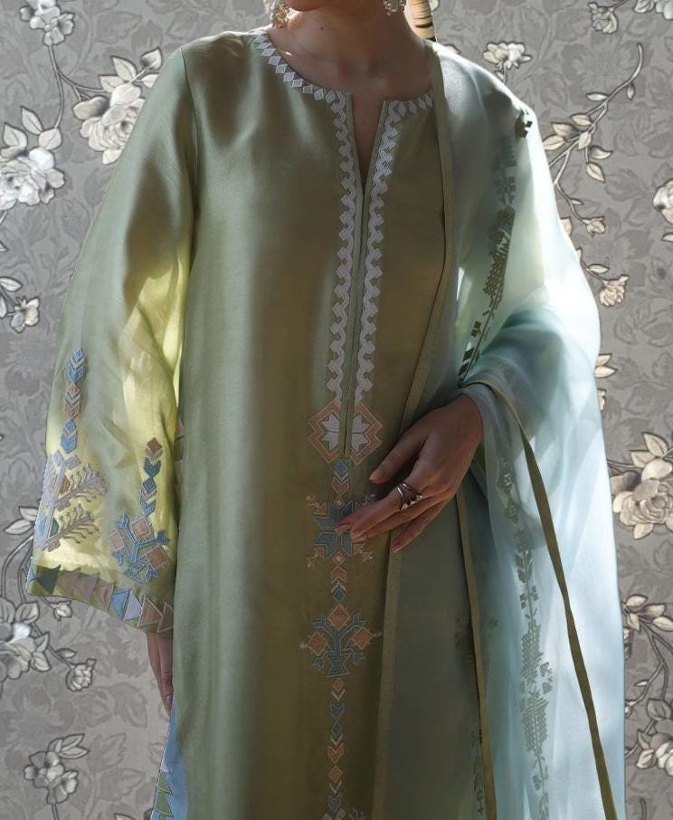 A person wearing a green dress with intricate embroidered patterns and designs, standing against a floral background.