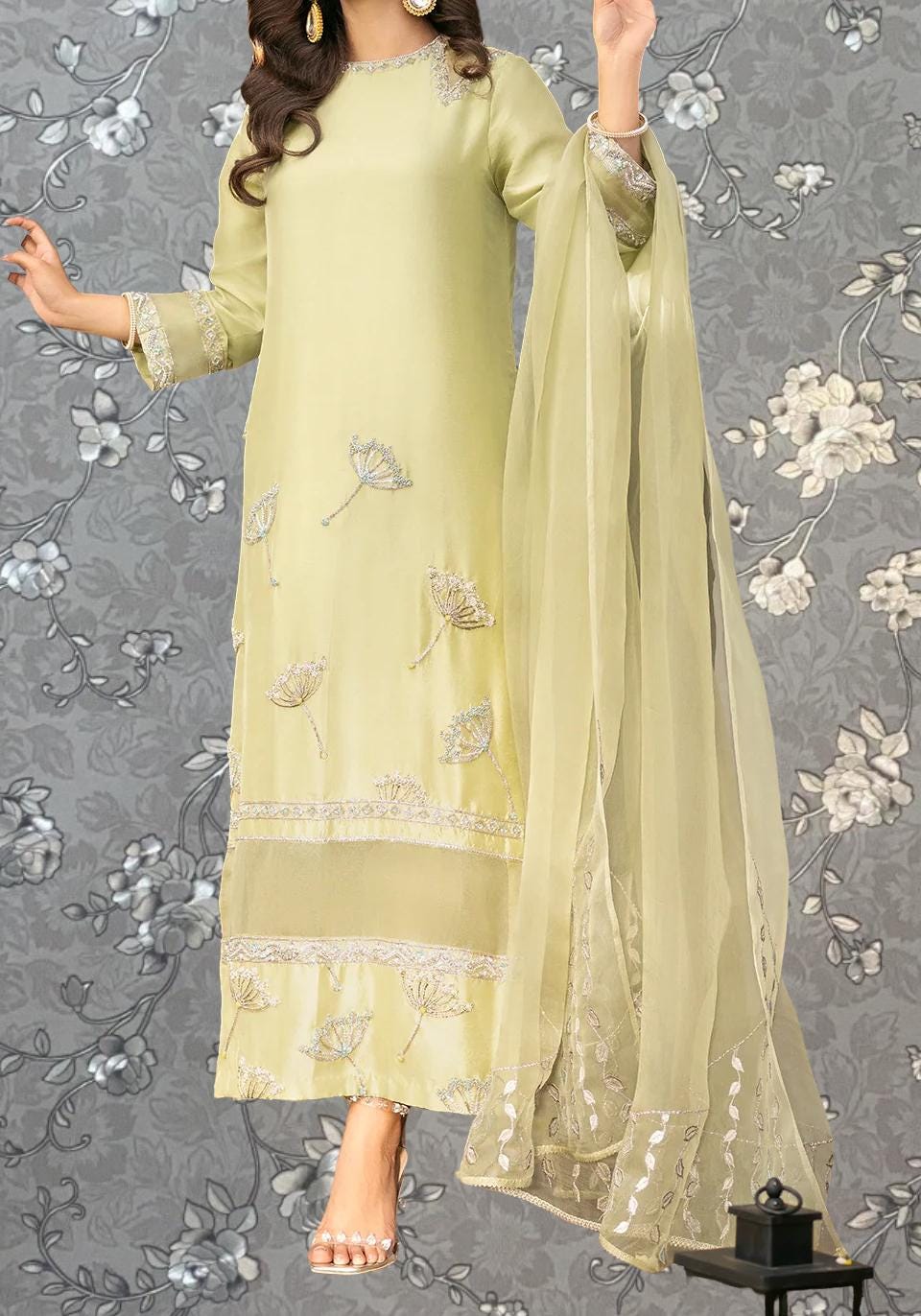 A woman wearing a long, yellow dress with intricate embroidered designs on it, standing against a gray floral background.