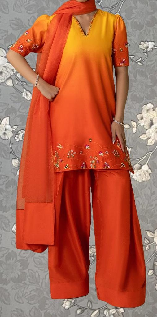 A person wearing a vibrant orange and yellow dress with floral embroidery, paired with matching orange pants, stands against a gray floral background.