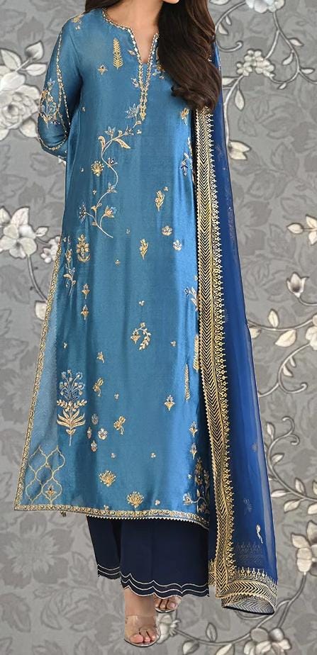 A woman wearing a blue and gold dress with intricate embroidery, standing against a gray floral background.
