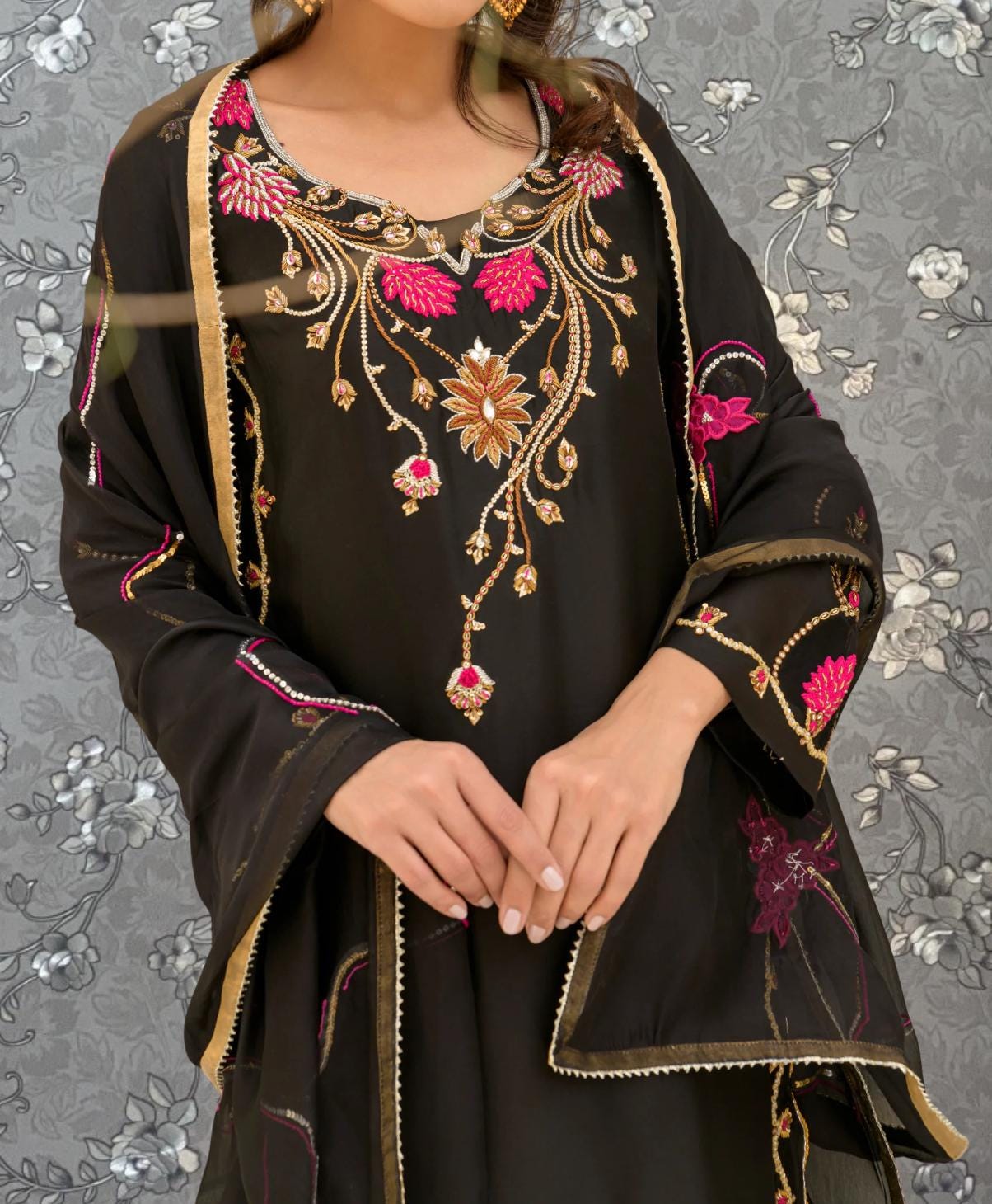 A woman wearing a black dress with intricate floral embroidery and a matching shawl stands against a gray floral background.