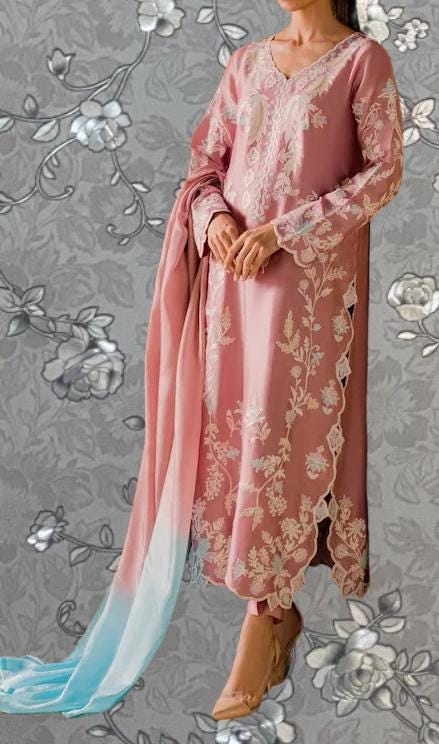 A woman wearing a pink dress with intricate floral embroidery stands against a gray floral background.