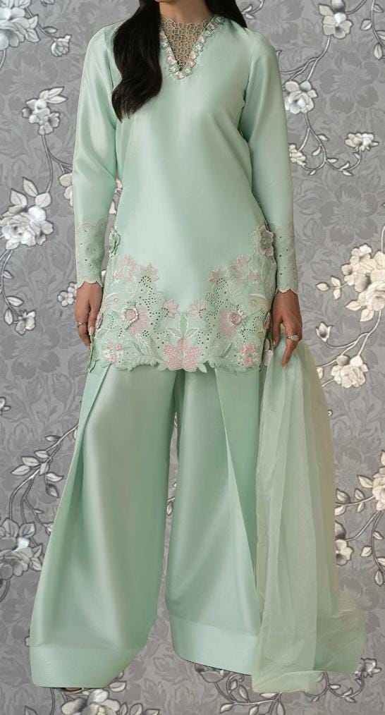 A woman wearing a mint green dress with floral embroidery and a matching green skirt, standing against a gray floral background.