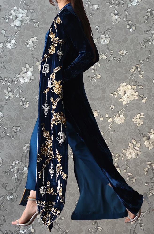 A woman wearing a long, blue velvet coat with gold embroidery walks against a gray floral background.