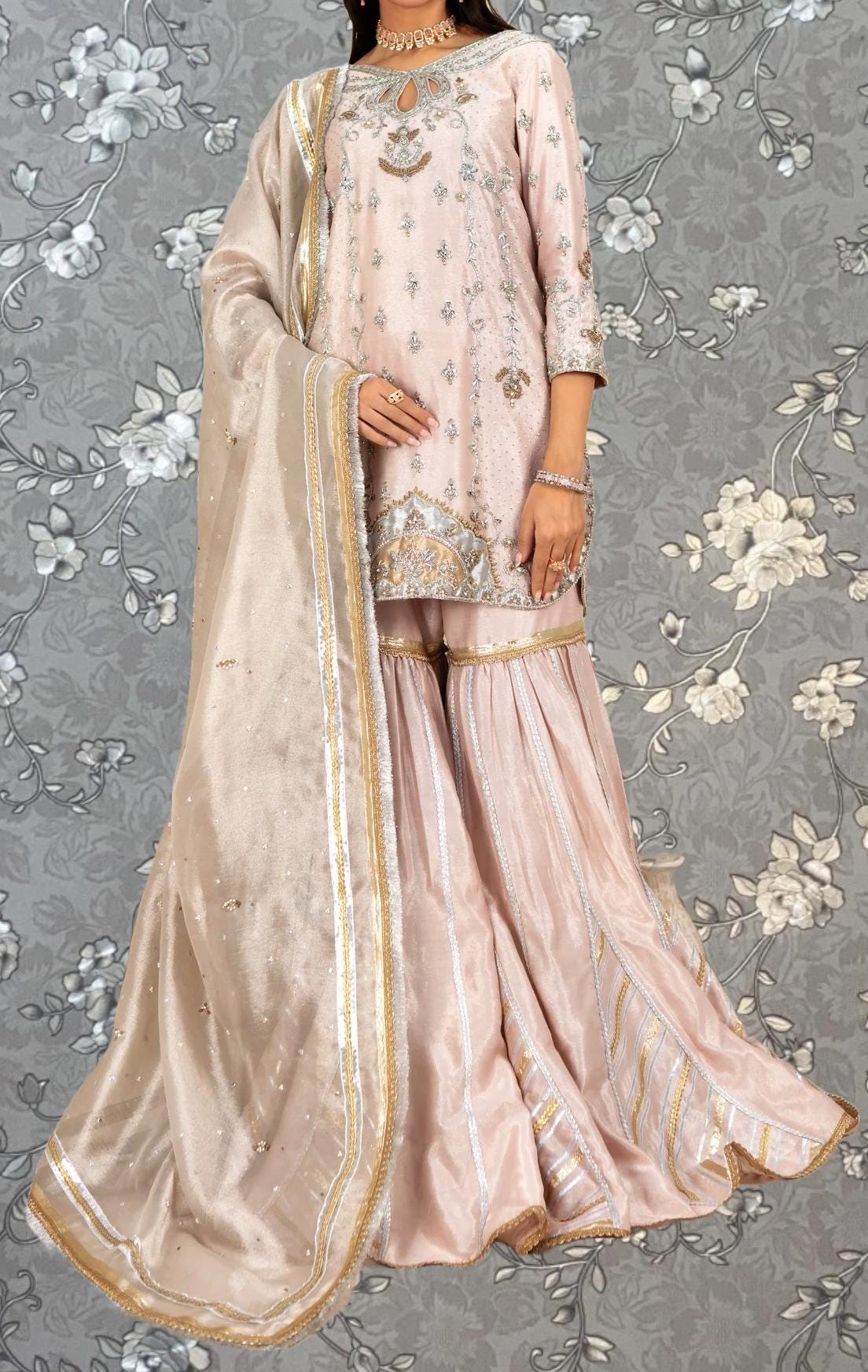 A woman wearing a pink and gold dress with a matching shawl, standing against a gray floral background.