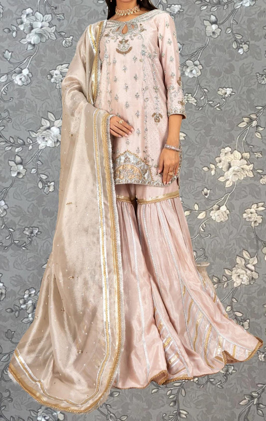 A woman wearing a pink and gold dress with a matching shawl, standing against a gray floral background.