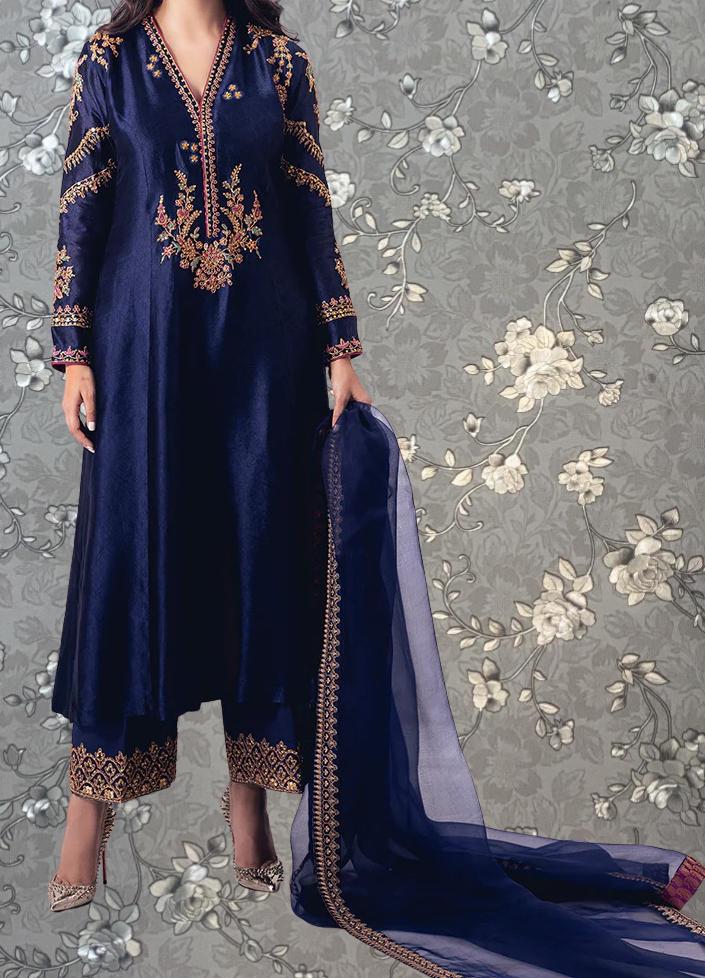 A woman wearing a long navy blue dress with gold embroidery and a matching blue shawl stands against a gray floral background.