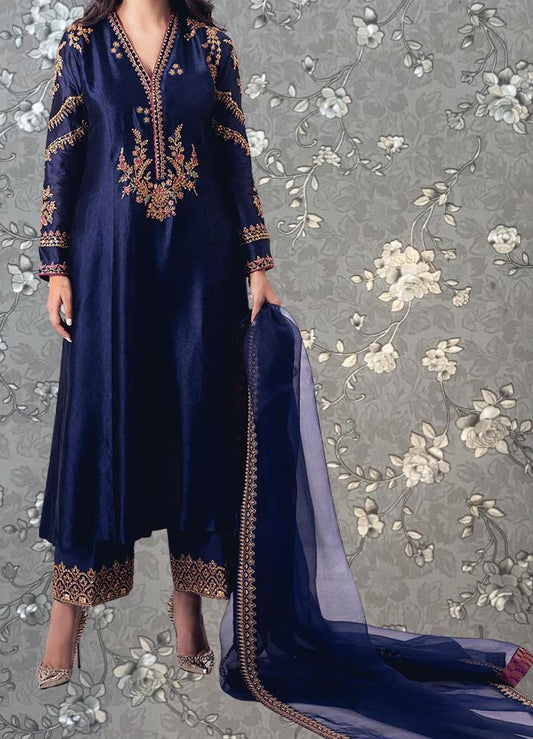 A woman wearing a long navy blue dress with gold embroidery and a matching blue shawl stands against a gray floral background.