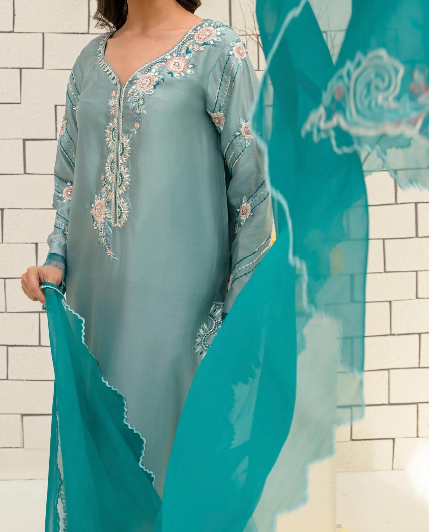 A woman wearing a light blue dress with intricate floral embroidery stands in front of a white brick wall.