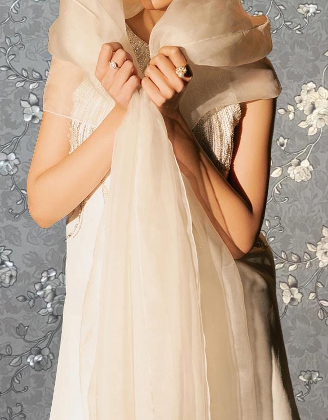 A woman in a white dress stands against a floral background, holding a sheer white fabric in her hands.