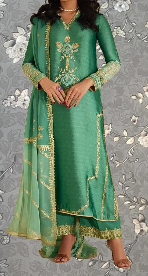 A woman wearing a green and gold dress with intricate embroidery and gold trim, standing against a gray floral background.