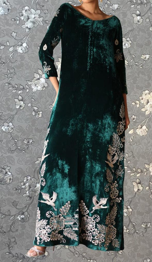 A woman wearing a long, green velvet dress with intricate gold embroidery on the sleeves and hem.