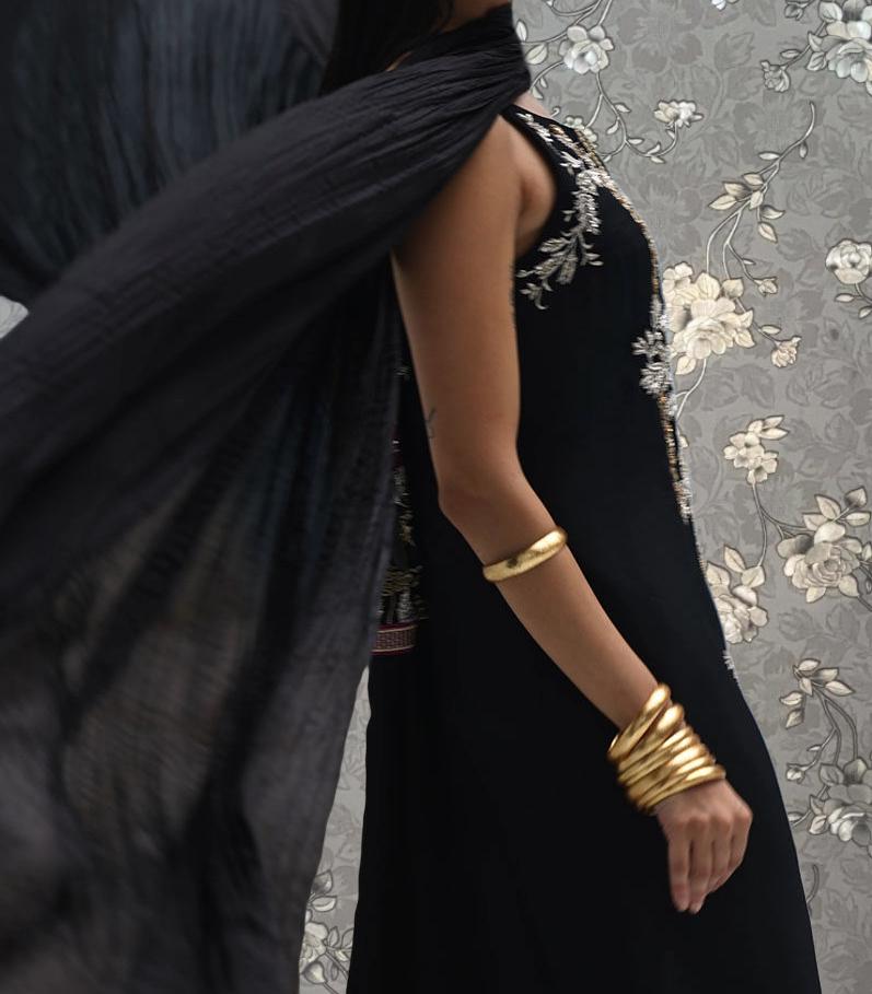 A woman wearing a black dress with gold accents and bracelets is standing in front of a floral patterned wall.