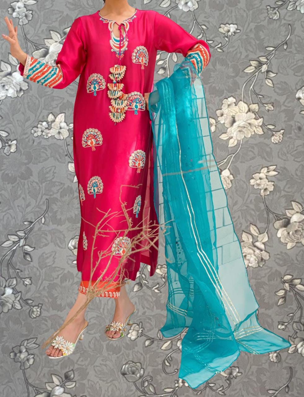 A person wearing a vibrant red dress with intricate peacock designs and a matching turquoise shawl stands against a gray floral background.
