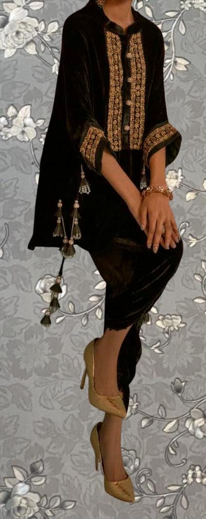 A person wearing a black velvet dress with gold embroidery and tassels, paired with gold high heels, stands against a gray floral background.