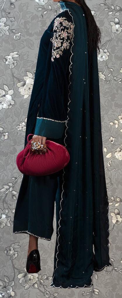 A person wearing a long, dark blue dress with intricate embroidery and a matching shawl, holding a red clutch purse, standing against a floral patterned background.