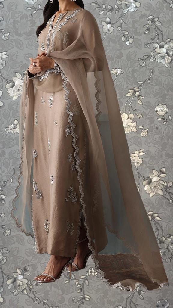 A woman wearing a long, beige dress with intricate embroidery and a matching shawl, standing against a gray floral background.