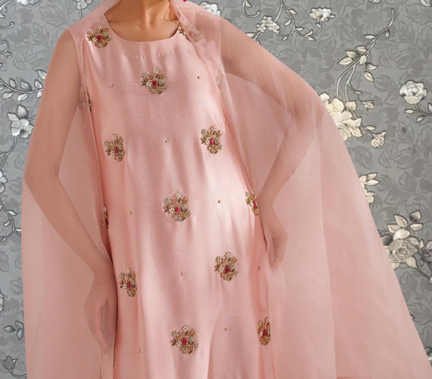 A close-up of a woman wearing a pink dress with gold embroidery, standing against a gray floral background.